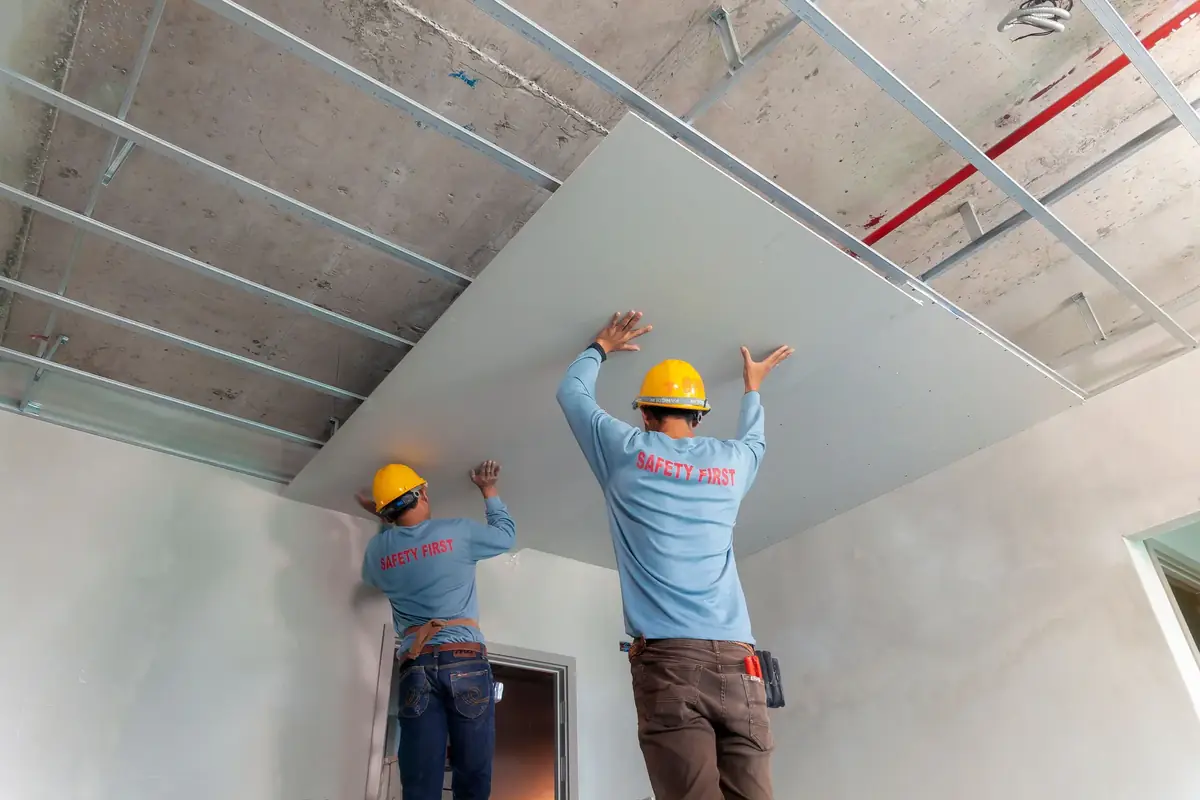 Two installers in hard hats lifting a drywall sheet